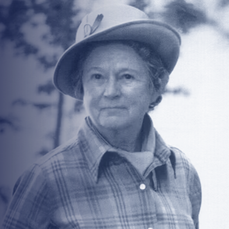 An older woman wearing a wide-brimmed hat and a plaid button-up shirt, standing outdoors with blurred trees in the background.